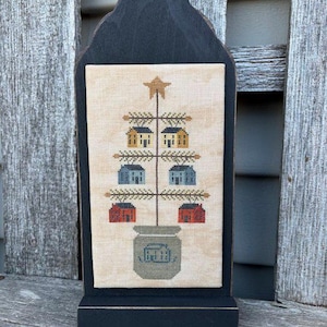 Pode incluir: Um desenho de ponto cruz de uma árvore de Natal com casas como ornamentos. A árvore está em um vaso com a palavra "Alegria" nele. O desenho é emoldurado em uma moldura de madeira preta.