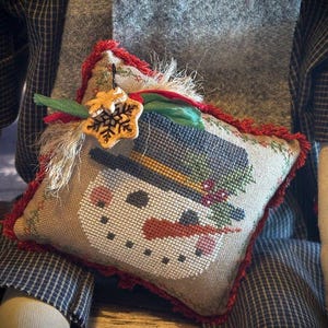 Peut inclure: Un petit coussin carré brodé au point de croix avec un bonhomme de neige. Le bonhomme de neige porte un chapeau haut de forme noir, un nez en carotte et une écharpe rouge. Le coussin est bordé de franges rouges et orné d'un flocon de neige.