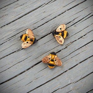 May include: Three decorative bee-shaped embellishments. Each bee features a black and yellow striped body, tan wings, and a black head. The bees are arranged on a gray, weathered wooden surface.