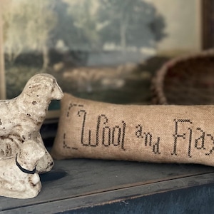 May include: A small, white, ceramic lamb figurine with a lamb laying on its back next to it. A tan pillow with the text "Wool and Flax" embroidered in black thread is in the background.