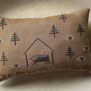 Puede incluir: Una almohada de color beige con un diseño de punto de cruz que representa una escena de la natividad con un pesebre, una estrella y árboles. El texto "Our Dear Savior's Birth" está bordado en la parte inferior de la almohada.