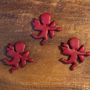 May include: Three red cupid figurines with wings and bows and arrows are arranged on a wooden surface. The figurines are in a running pose, with their bows drawn. The background is a warm, brown wood grain.