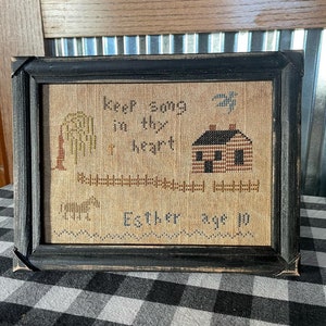 Könnte beinhalten: Ein gerahmter Kreuzstich-Sampler mit einem schwarzen Rahmen und einem beigefarbenen Hintergrund. Der Sampler zeigt ein Haus, einen Baum, einen Vogel und den Text "Keep song in thy heart Esther age 10".