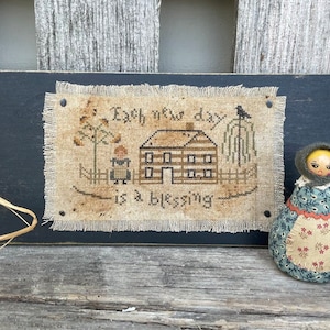Puede incluir: Un letrero de madera negro con un diseño de punto de cruz que dice "Cada nuevo día es una bendición". El diseño presenta una casa, un árbol y un pájaro. Una pequeña muñeca con la cara amarilla y un vestido azul está sentada a la derecha del letrero.