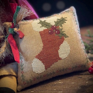 Peut inclure: Un petit coussin carré beige représentant une chaussette de Noël brodée au point de croix. La chaussette est rouge avec des accents blancs, surmontée de houx vert et de baies rouges. Le coussin est bordé de coutures vertes et rouges et noué avec des rubans rouges et verts.