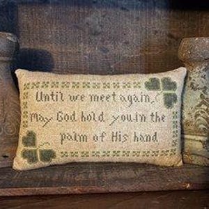 Puede incluir: Una almohada beige con tréboles verdes bordados. La almohada tiene el texto "Until we meet again, May God hold you in the palm of His hand" bordado.