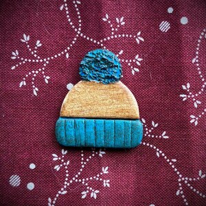 May include: A wooden pin shaped like a winter hat. The hat has a blue pom-pom, a tan mid-section, and a blue ribbed brim. The pin is set against a burgundy fabric with a white floral pattern.