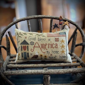 Op de afbeelding: Een rechthoekig, beige, geborduurd kussen met patriottische afbeeldingen, waaronder een Amerikaanse vlag, een huis en de woorden "God bless America my home sweet home". Het kussen is versierd met stervormige spelden en ligt op een rustieke houten stoel.