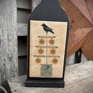 May include: A black wooden framed decorative piece with a stitched design. The design features a black crow perched on a floral stem with yellow flowers. The stem is in a jar with a bird design.