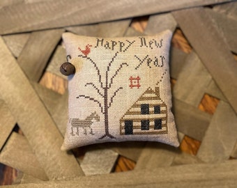 May include: A beige pillow with a cross-stitch design featuring a house, a tree, a horse, and a bird. The pillow has the text "Happy New Year" stitched on it.