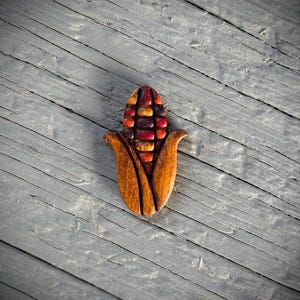 May include: A wooden corn-shaped decorative item. The corn features a colorful design with red, orange, and yellow kernels, and brown husks. The item is placed on a gray wooden surface.
