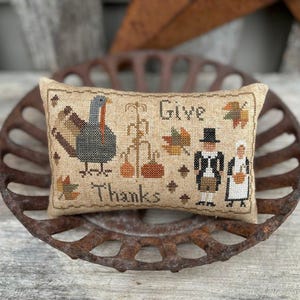 Op de afbeelding: Een beige kussen met een kruissteekontwerp met een kalkoen, pompoenen, maïsstengels en twee figuren in pelgrimkleding. De tekst "Give Thanks" is op het kussen geborduurd.