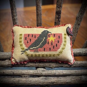 May include: A rectangular fabric pincushion featuring a cross-stitch design of a black bird perched on a slice of watermelon with a yellow star. The cushion has a textured, rust-colored trim and a small, dark button attached to the upper right corner.