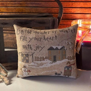 May include: A decorative pillow with a winter scene and the text "Let the wonder of winter fill your heart with joy". The pillow is on a wooden surface with a candle in the background.