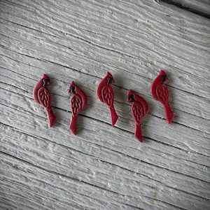 May include: Five red cardinal bird-shaped ornaments with black accents are arranged on a weathered, gray wooden surface. The ornaments have detailed wing and tail designs, and are evenly spaced.