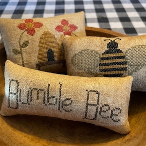 Peut inclure: Trois petits coussins brodés dans un bol en bois. Les coussins sont en tissu de jute et présentent des motifs sur le thème des abeilles. Un coussin a une ruche, un autre une abeille et le dernier dit "Bumble Bee".