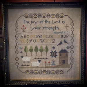 May include: Framed cross-stitch artwork featuring the text "The joy of the Lord is your strength." and "Nehemiah 8:10." The design includes trees, sheep, a house, and a star. The piece is framed in a dark, ornate frame.