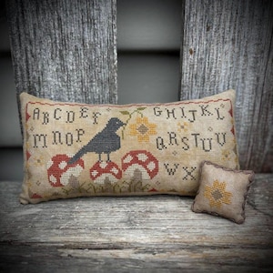 Puede incluir: Un cojín rectangular beige con bordado de punto de cruz. Presenta un cuervo negro posado sobre setas rojas y blancas, un borde alfabético y detalles florales. También hay un cojín cuadrado más pequeño con un diseño de flor amarilla.