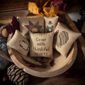 May include: A collection of small, burlap-textured pillows with embroidered designs, including a star, a pilgrim, a turkey, and a pumpkin. One pillow reads "Come with thankful hearts." The pillows are arranged in a wooden bowl with fall foliage.