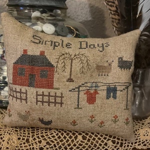 May include: A beige pillow with a cross-stitch design depicting a red house with a black roof, a fence, a tree, and clothes hanging on a clothesline. The text "Simple Days" is stitched in brown thread on the pillow.