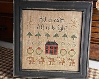 May include: A framed cross-stitch sampler with a beige background featuring a red house, green trees, and the text "All is calm, All is bright".