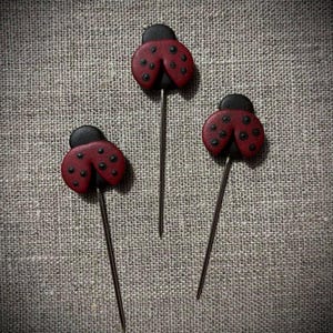 May include: Three decorative ladybug pins. Each pin features a red ladybug with black spots and a black head. The ladybugs are attached to long, thin, silver-colored pins. The pins are displayed on a textured, gray background.