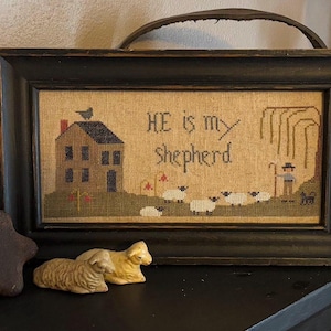 Peut inclure: Une broderie au point de croix encadrée avec un cadre noir et un fond beige. La broderie représente une petite maison, un berger et des moutons. Le texte "HE is my shepherd" est brodé au centre de l'image.