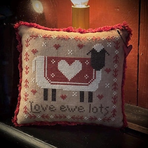 May include: A square, cross-stitched pillow with a sheep design. The sheep has a red blanket with a white heart. The pillow has a red fringe and the words "love ewe lots".