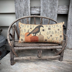 Puede incluir: Un cojín rectangular decorativo con un diseño de cuervo y araña, apoyado en un banco de madera rústico. El cojín presenta un cuervo posado sobre tres calabazas, con un borde alfabético. La paleta de colores incluye beige, naranja, marrón y negro.