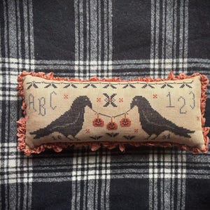 May include: A rectangular beige fabric pillow with a cross-stitch design featuring two black crows holding a string of three orange pumpkins. The letters ABC and numbers 123 are stitched in black. The pillow has a ruffled orange fabric border.