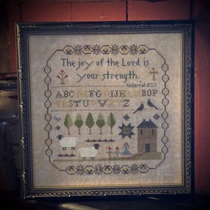 Puede incluir: Obra de arte en punto de cruz enmarcada con el texto "The joy of the Lord is your strength." y "Nehemiah 8:10." El diseño incluye árboles, ovejas, una casa y una estrella, todo en tonos apagados. El marco es oscuro.