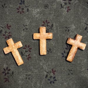 May include: Three small, wooden crosses with a light brown finish are arranged on a dark green fabric background with a floral pattern. The crosses are evenly spaced and slightly angled.