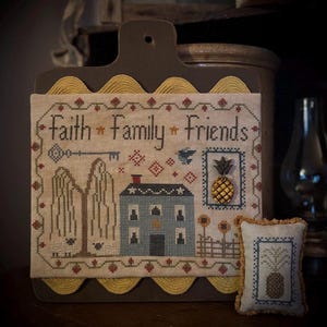Puede incluir: Un muestrario de punto de cruz enmarcado con las palabras "Faith Family Friends" sobre una casa azul, una llave y una piña. Un cojín más pequeño a juego con un diseño de piña está a la derecha. El muestrario está montado en una tabla de madera marrón oscuro.