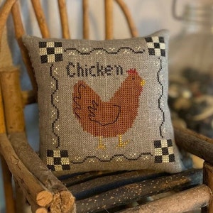 May include: A gray and white pillow with a brown chicken embroidered on it. The pillow says "Chicken" in black lettering. The pillow is sitting on a wooden chair.