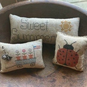 Peut inclure: Trois housses de coussin brodées sur le thème de l'été. Un coussin porte l'inscription "Sweet Summer" avec un motif de soleil. Un autre coussin représente une coccinelle rouge. Le troisième coussin a un petit chariot avec des fleurs.