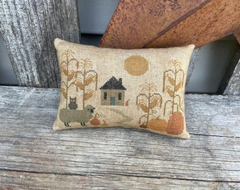 May include: A beige pillow with an embroidered autumn scene. The scene includes a small house, a black cat, a sheep, corn stalks, pumpkins, and a sun.