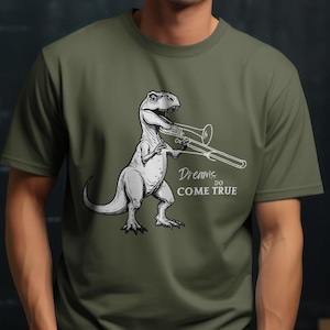 以下が含まれることがあります： トロンボーンを演奏する恐竜の白いグラフィックが描かれた緑色のTシャツ。恐竜の下には「Dreams do come true」という文字が印刷されています。
