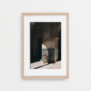 May include: A wooden chair sits in a sunlit courtyard with intricate tilework and arched doorways. The chair is positioned in the shadow of a pillar, creating a dramatic contrast of light and dark.