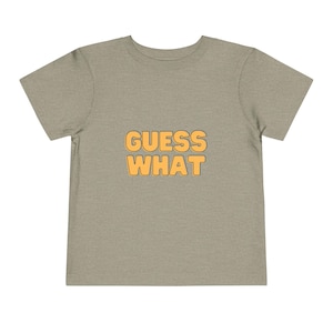 May include: A toddler t-shirt in a neutral tan color. The shirt features the text "GUESS WHAT" in a bold, orange font. The text is centered on the front of the shirt.