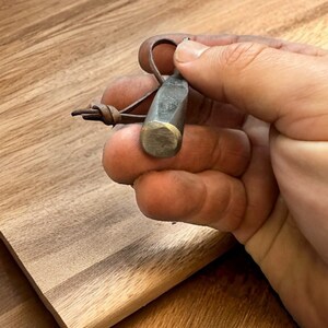 Close-up of the pipe tamper held between fingers, with its flat tamping face toward the camera and the twisted shaft and top loop behind it.