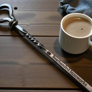 May include: A hand-forged metal bottle opener with a twisted handle and loop. The opener is engraved with "Bearhanded Ironworks." A white mug of coffee sits next to the opener on a wooden surface.