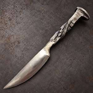 Puede incluir: Un cuchillo de hoja fija forjado a partir de un clavo de vía férrea, con una hoja de acero pulido de tono plateado y un mango texturizado de color gris oscuro con un diseño de metal retorcido que conserva la forma original.