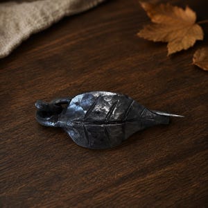 May include: A dark gray, hand-forged metal brooch shaped like a leaf. The surface features a hammered, textured finish with deep, incised lines representing veins. A pointed metal pin extends from the right side of the leaf-shaped body.