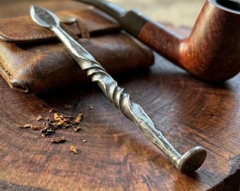 May include: A hand-forged metal pipe tamper featuring a twisted handle, a flat circular base, and a pointed tip. The tool is positioned alongside a brown leather tobacco pouch and a wooden smoking pipe on a dark, circular wooden surface.