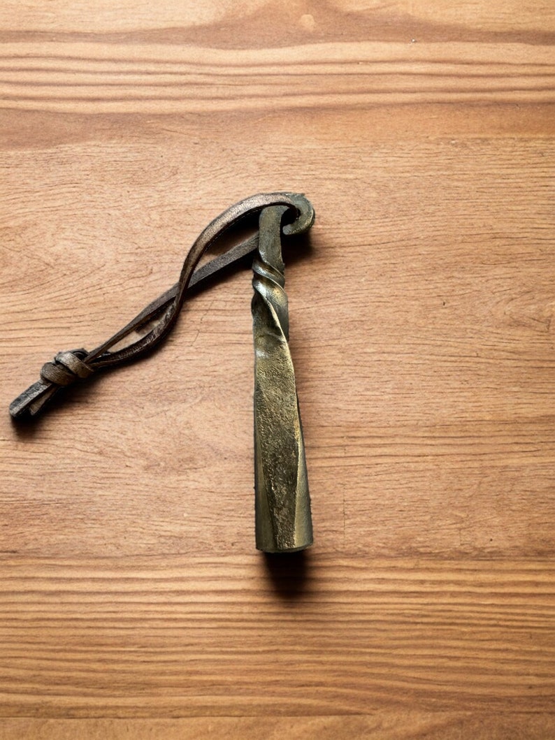 Hand-forged steel pipe tamper on a wooden surface, showing its twisted shaft, curled top and attached brown loop.