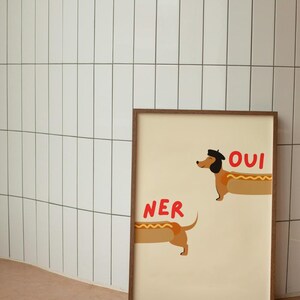 K&ouml;nnte beinhalten: Gerahmter Kunstdruck mit zwei Dackeln als Hotdogs. Einer tr&auml;gt einen Hut, dar&uuml;ber steht "OUI". &Uuml;ber dem anderen steht "NER". Creme Hintergrund, brauner Holzrahmen.