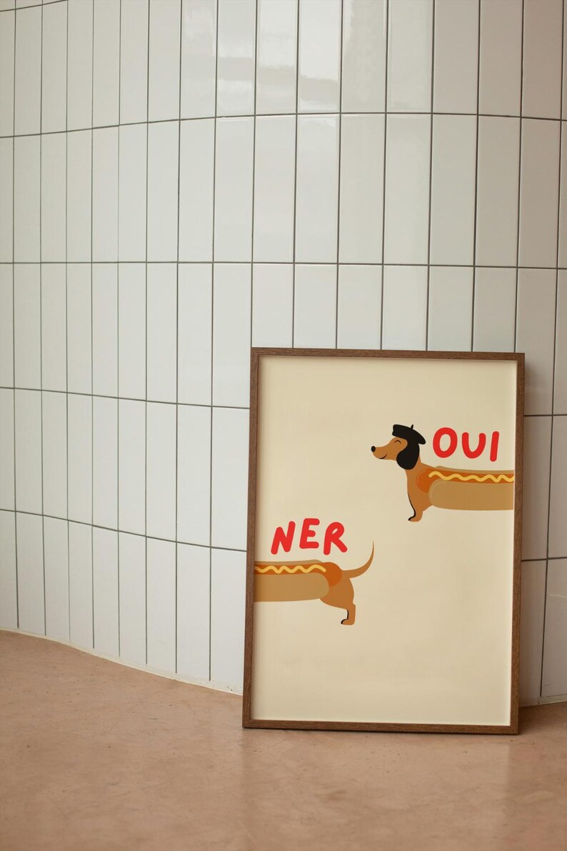 K&ouml;nnte beinhalten: Gerahmter Kunstdruck mit zwei Dackeln als Hotdogs. Einer tr&auml;gt einen Hut, dar&uuml;ber steht "OUI". &Uuml;ber dem anderen steht "NER". Creme Hintergrund, brauner Holzrahmen.