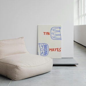 Pu&ograve; includere: Un pouf beige &egrave; posizionato accanto a una stampa su tela. La stampa presenta un'illustrazione blu e rossa di sardine in scatola con le parole "TIN MATES" scritte in rosso e blu.