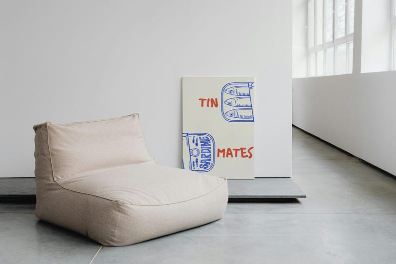 Pu&ograve; includere: Un pouf beige &egrave; posizionato accanto a una stampa su tela. La stampa presenta un'illustrazione blu e rossa di sardine in scatola con le parole "TIN MATES" scritte in rosso e blu.