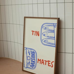 Pu&ograve; includere: Una stampa incorniciata con un'illustrazione di sardine in scatola. Il design utilizza blu e rosso, con le parole "TIN MATES". La cornice &egrave; marrone. L'opera d'arte raffigura sardine in scatola, con un carattere giocoso.